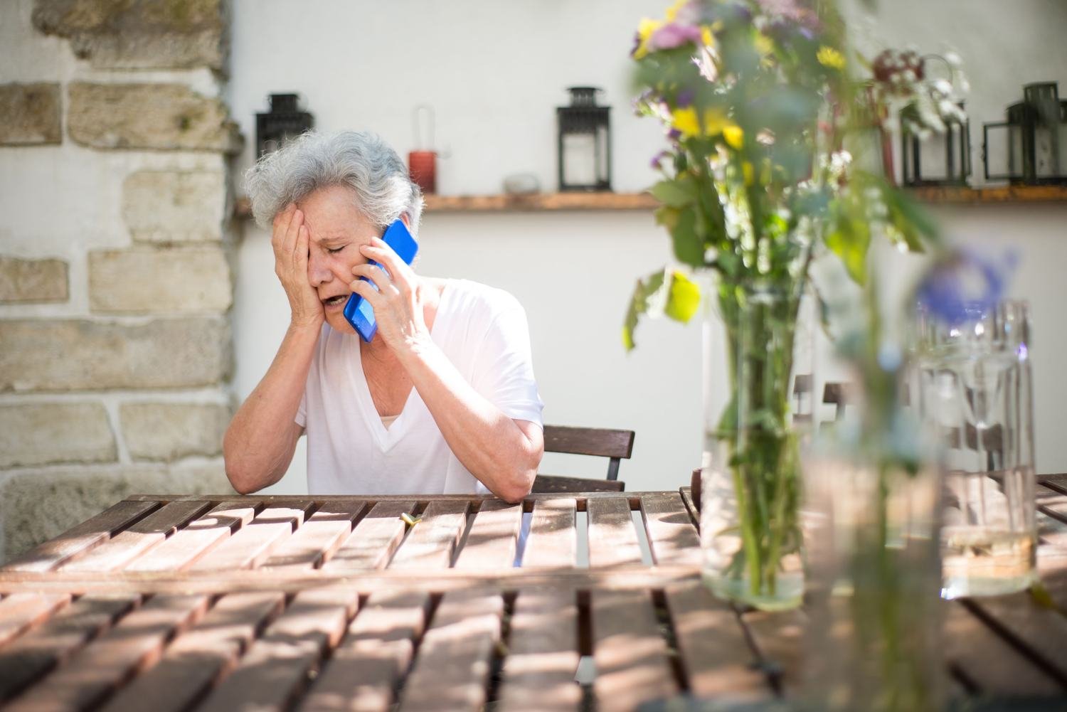 An older woman on the phone at an outdoor table, in distress from tooth pain